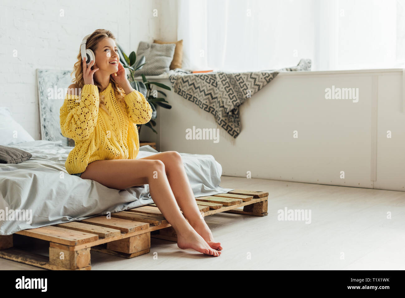 Beautiful smiling young woman in headphones assis sur le lit et écoute la musique à la maison with copy space Banque D'Images