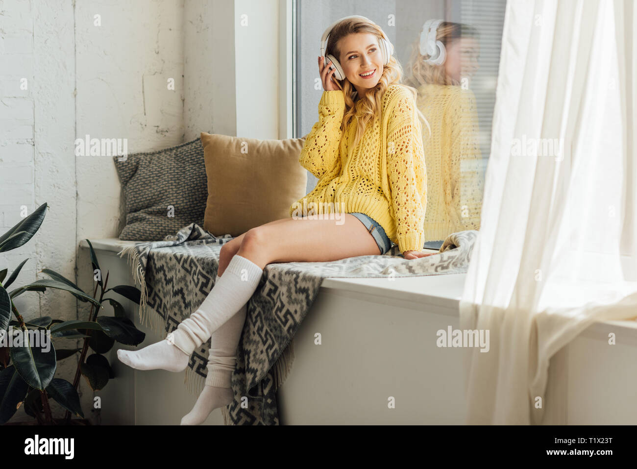 Beautiful smiling young woman in headphones assis sur un rebord de fenêtre et écouter la musique à la maison Banque D'Images