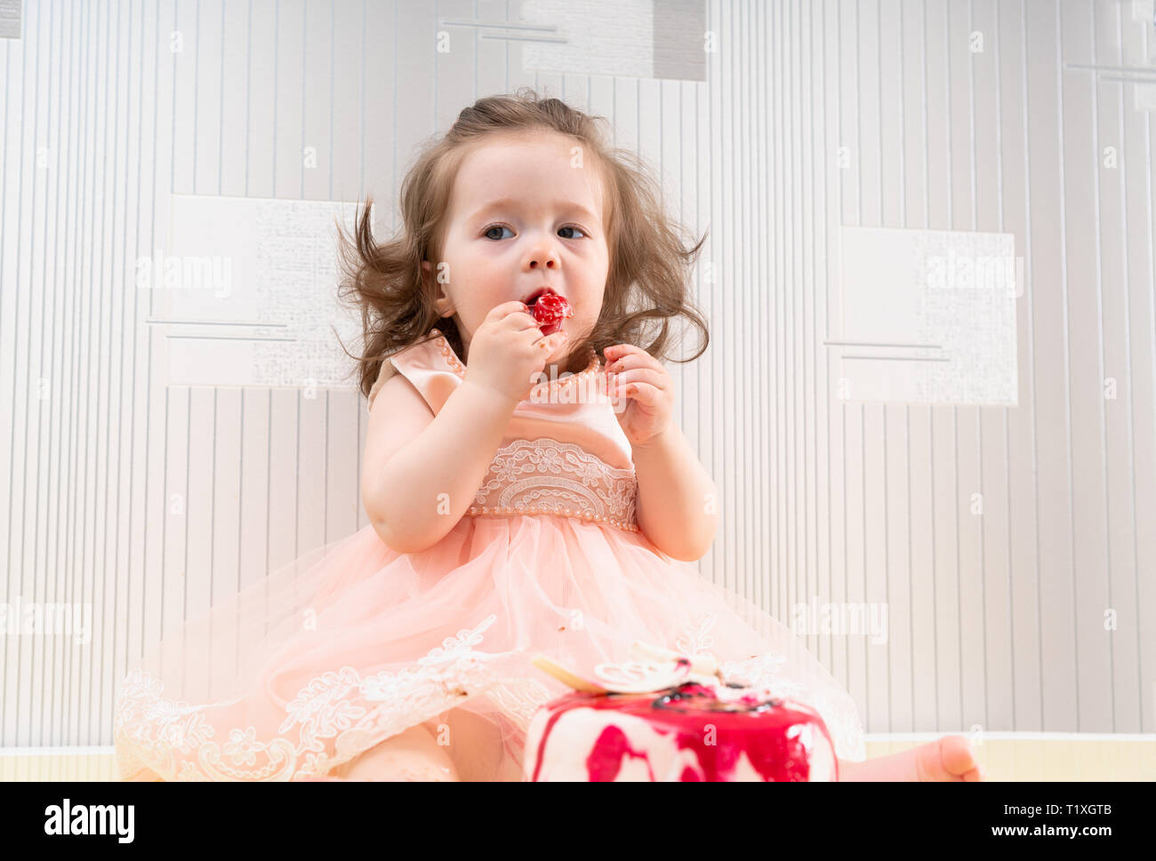 Petite Fille En Robe Rose Eating De Sweet Gateau Avec Glacage Rouge Assis Sur La Table Contre Le Mur D Argent Photo Stock Alamy