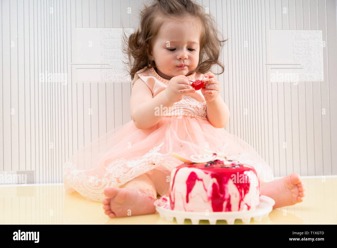 Petite Fille En Robe Rose De Manger Un Gateau Avec Ses Doigts Assis Sur La Surface De La Table Et En Regardant Un Morceau De Cerise Rouge Dans Ses Mains Photo Stock