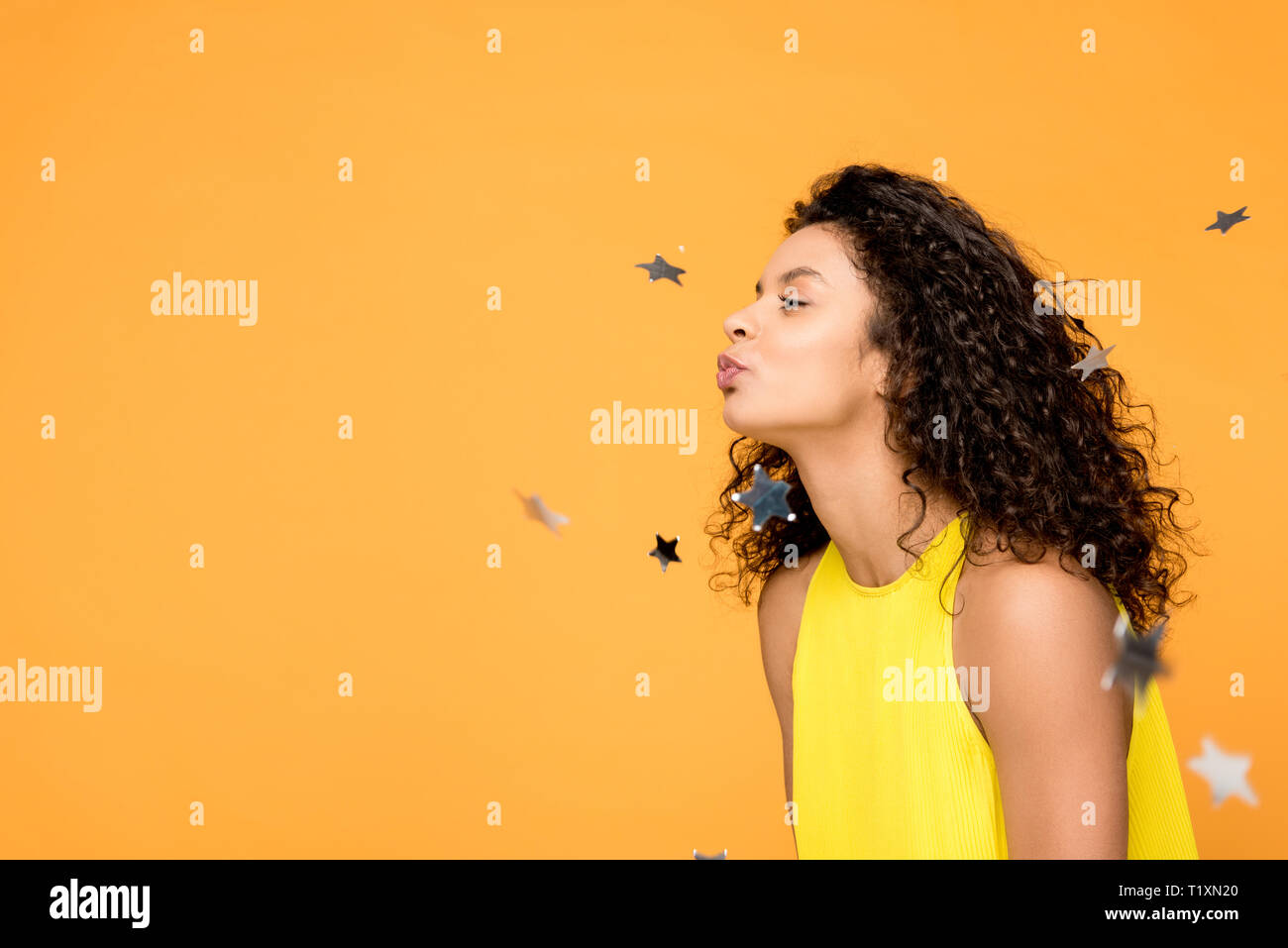 Heureux les african american girl robe jaune en soufflant près de confettis étoiles brillant isolé sur orange Banque D'Images