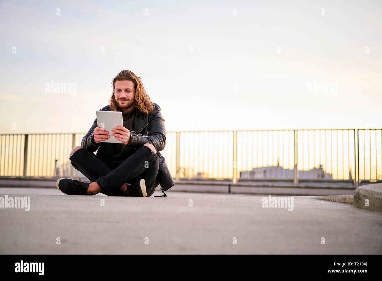 Portrait de jeune homme barbu assis sur niveau parking au crépuscule using digital tablet Banque D'Images