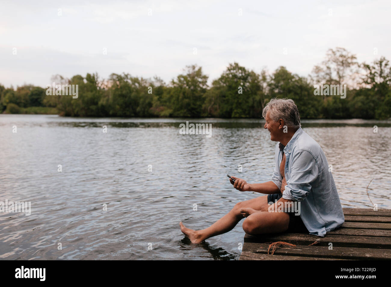 Happy senior man sitting on jetty at a lake with cell phone Banque D'Images