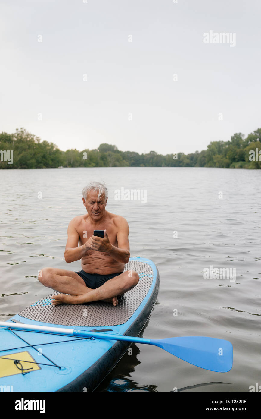 Senior man sitting on conseil SUP sur un lac using cell phone Banque D'Images