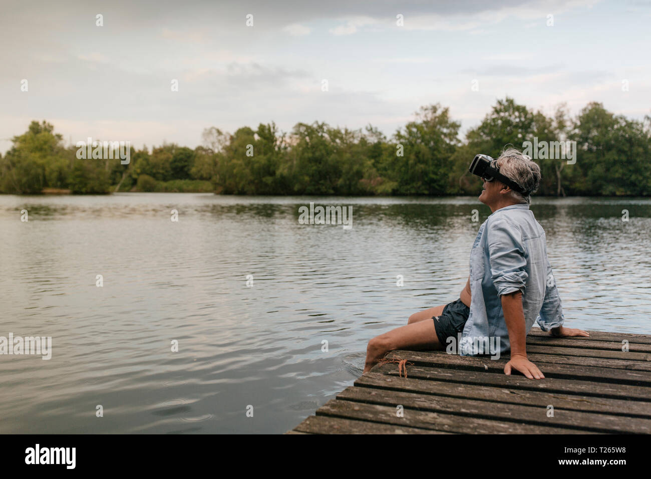 Senior man sitting on jetty at un lac portant des lunettes VR Banque D'Images