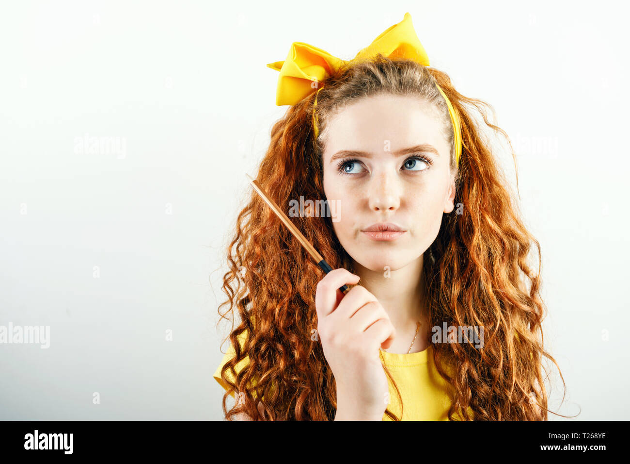 Pensive gingembre curly girl avec un arc jaune sur sa tête tenant un crayon et pensez à quelque chose en étant debout sur le fond blanc Banque D'Images