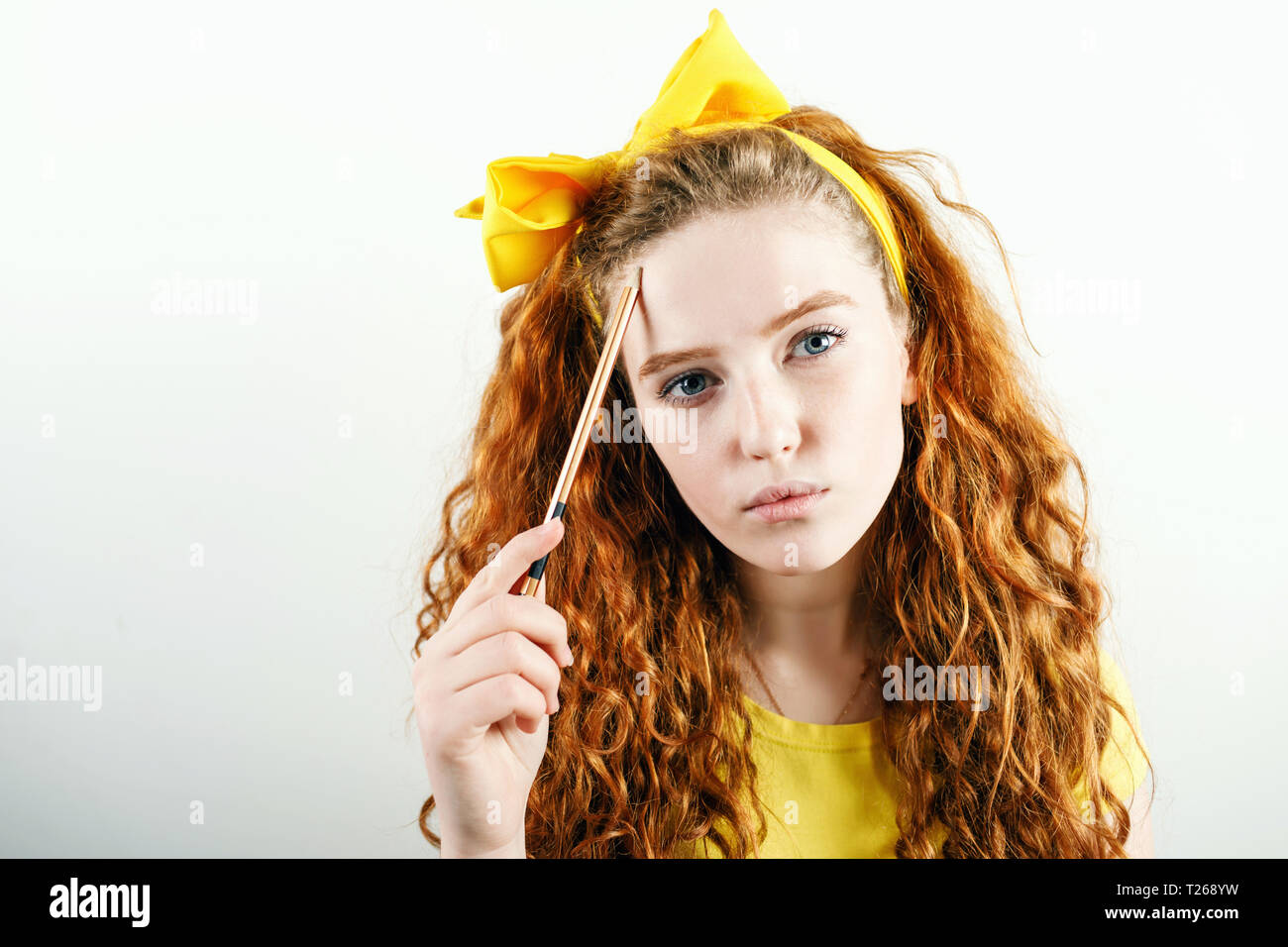 Pensive gingembre curly girl avec un arc jaune sur sa tête holding pencil près de son visage et de penser à quelque chose en étant debout sur le fond blanc. Banque D'Images
