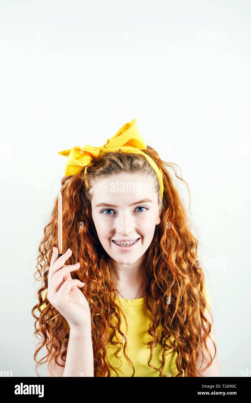 Cheerful curly ginger girl entre parenthèses avec un arc jaune sur sa tête debout sur le fond blanc et la tenue de crayon, avoir idée concept Banque D'Images
