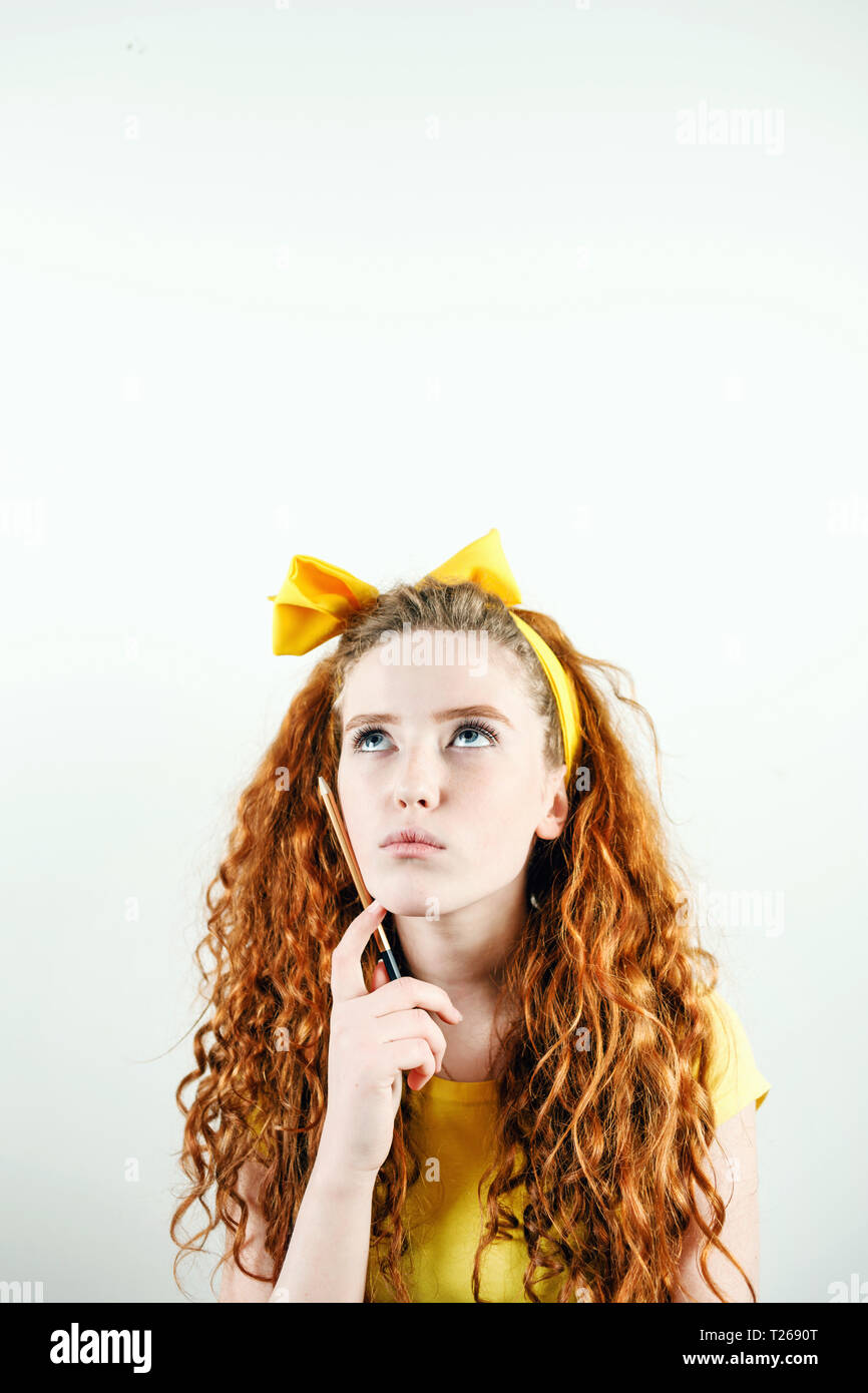 Pensive curly redhead girl avec un arc jaune sur sa tête debout sur le fond blanc, tenant un crayon et penser à quelque chose Banque D'Images