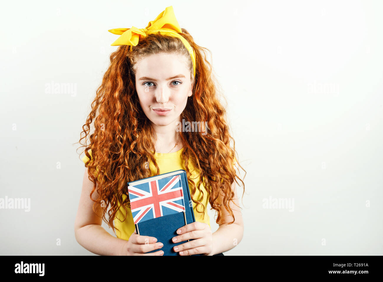 Fille rousse bouclée avec un arc jaune sur sa tête portant des T-shirt jaune holding book et drapeau britannique, debout sur le fond blanc Banque D'Images