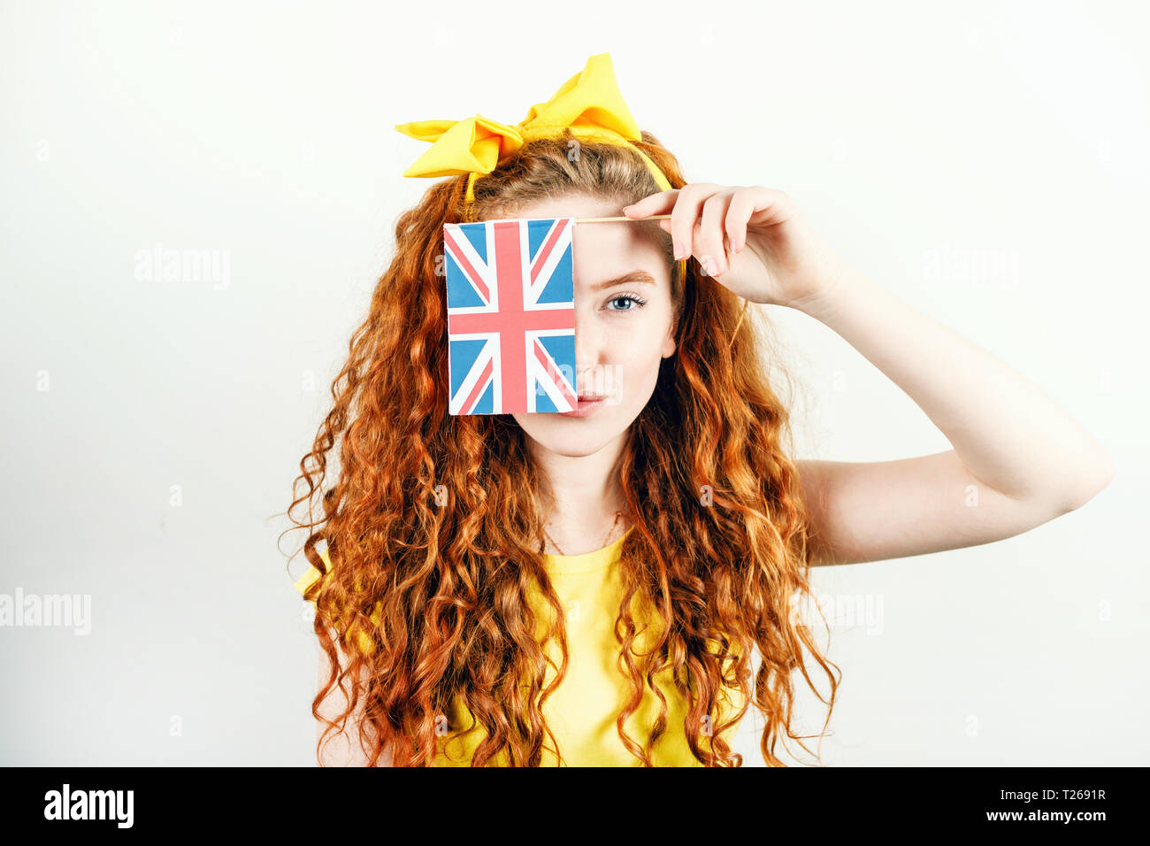 Fille rousse bouclée avec un arc jaune sur sa tête portant des T-shirt jaune dissimule son visage derrière le petit drapeau de l'Angleterre en étant debout sur le fond blanc. Banque D'Images