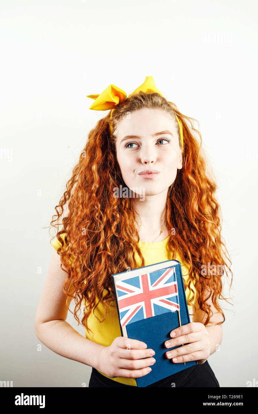 Fille rousse bouclée avec un arc jaune sur sa tête portant des T-shirt jaune holding book et drapeau britannique, à l'écart tout en se tenant sur la zone blanche Banque D'Images