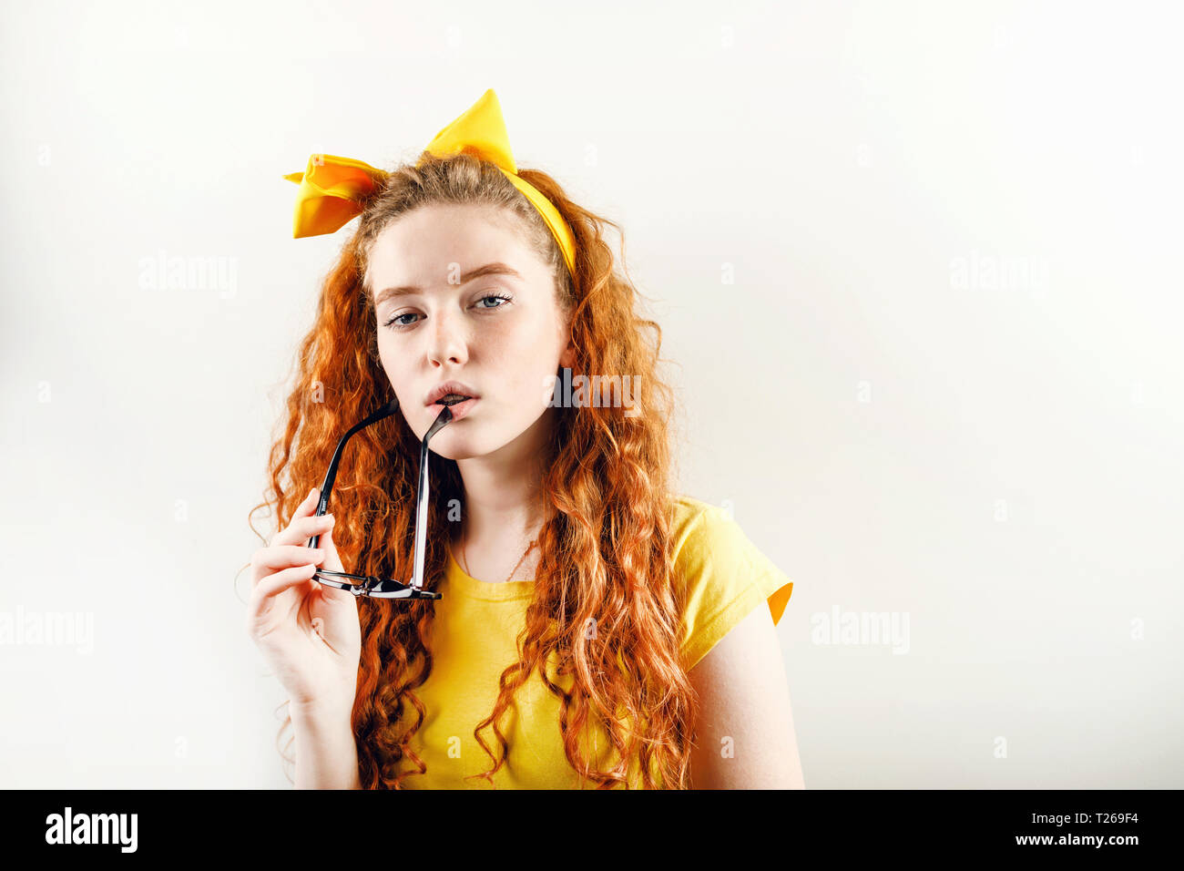 Pensive curly redhead girl avec un arc jaune sur sa tête portant des T-shirt jaune holding eyeglasses près de sa bouche et à la recherche de l'appareil photo Banque D'Images