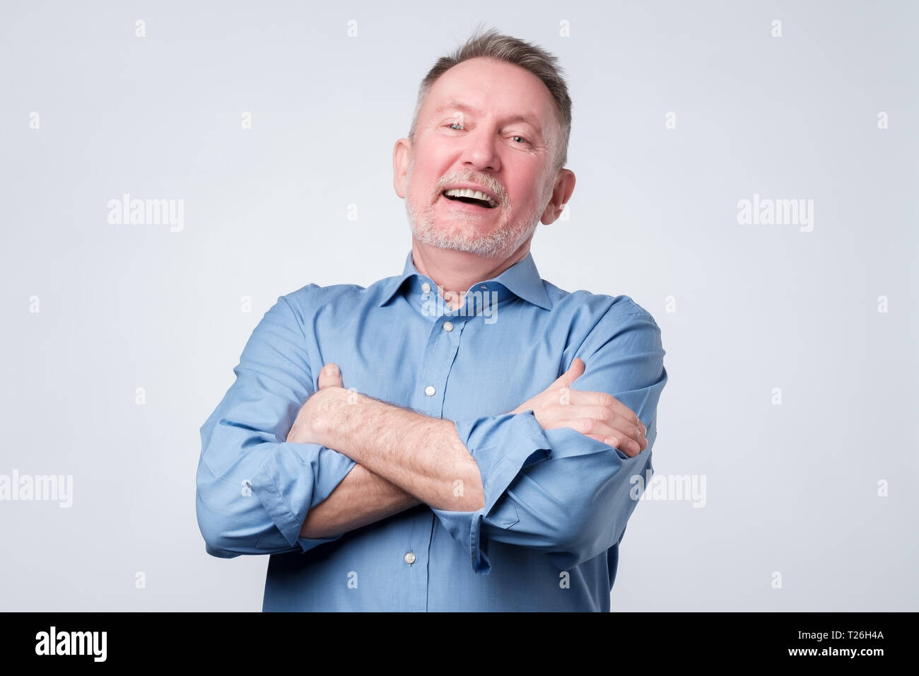 Cheerful senior man enjoying success. Vieil homme en chemise bleu bras repliables, détournant les yeux et sourit. La réussite de l'entreprise concept Banque D'Images