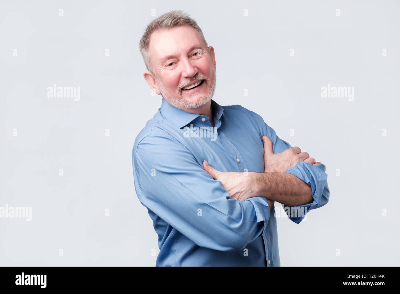 Cheerful senior man enjoying success. Vieil homme en chemise bleu bras repliables, détournant les yeux et sourit. La réussite de l'entreprise concept Banque D'Images