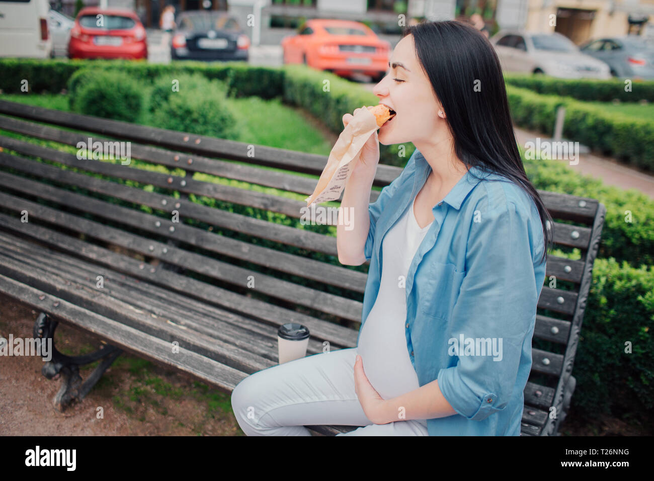 Portrait of a happy black hair et fière femme enceinte dans une ville à l'arrière-plan. Elle est assise sur un banc de la ville et de manger un gâteau ou un sandwich. Hung Banque D'Images