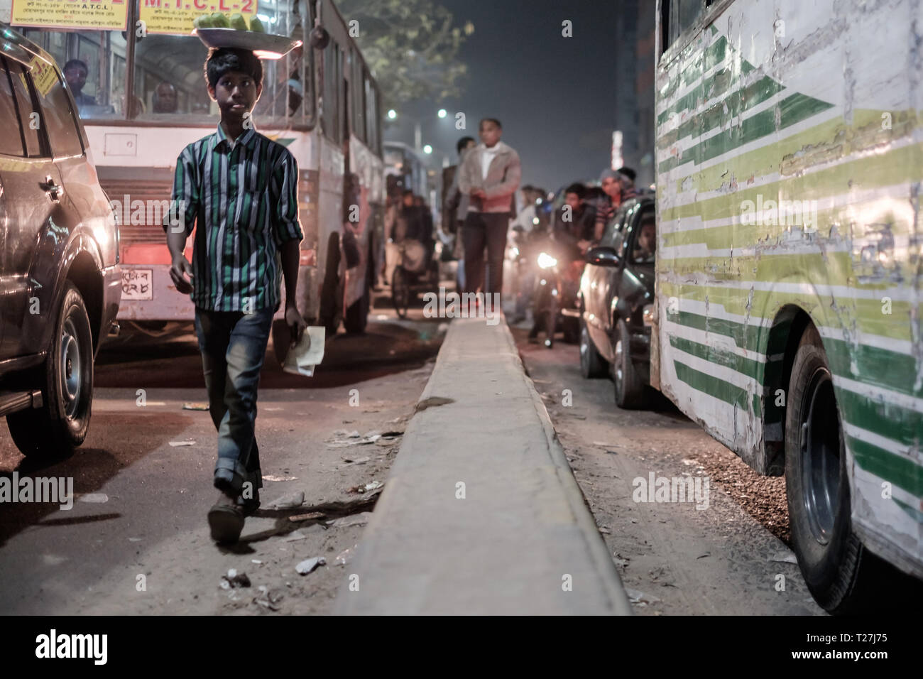 Vendeur de rue de l'adolescence de marcher au milieu de rue le soir à Dhaka, au Bangladesh. Voitures à attendre dans les embouteillages. Banque D'Images