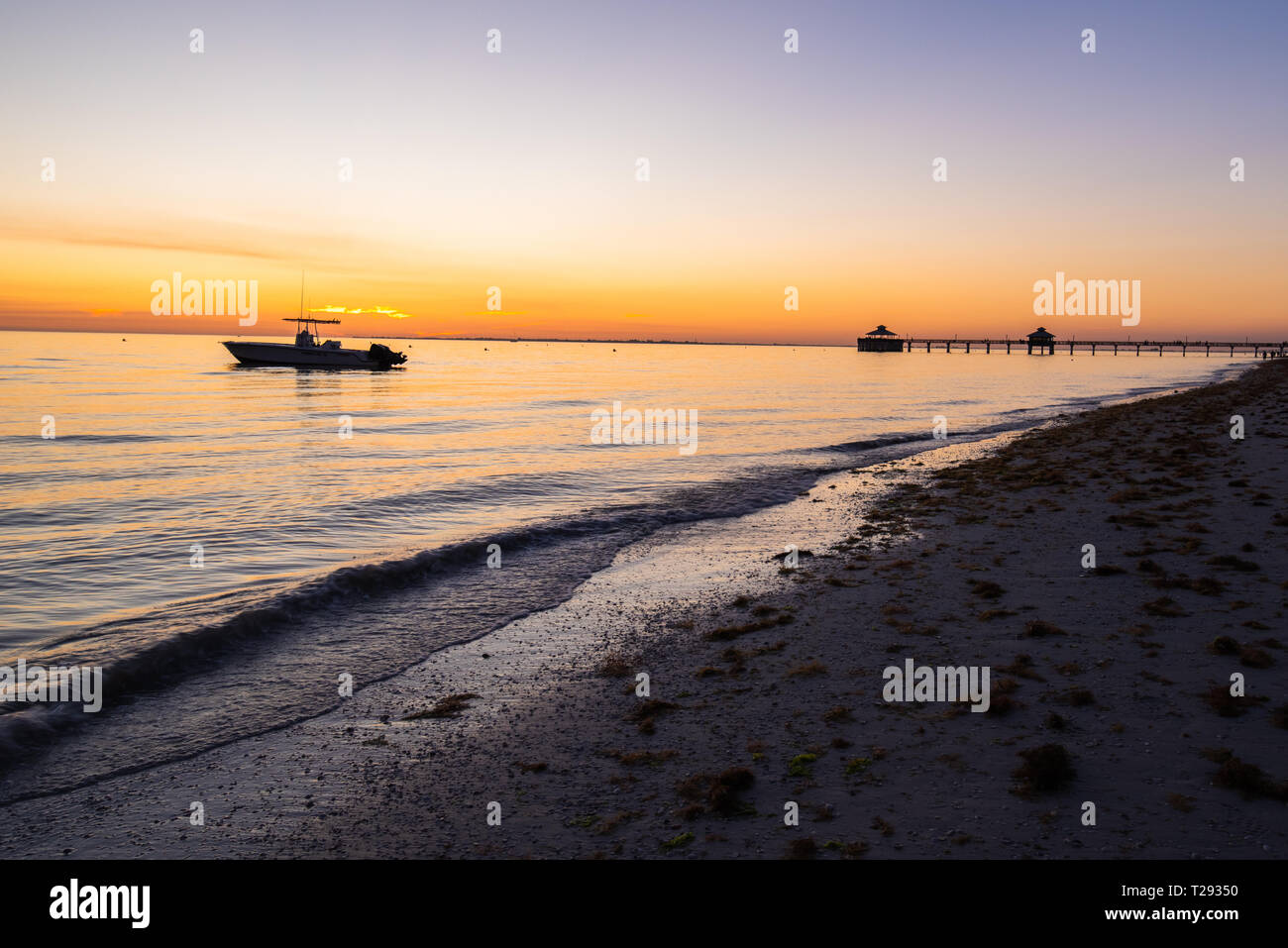 Un petit bateau de pêche est ancrée dans les eaux peu profondes de la plage de Fort Myers shore avec le quai de pêche dans l'arrière-plan et couleurs au coucher du soleil sur l'horizo Banque D'Images
