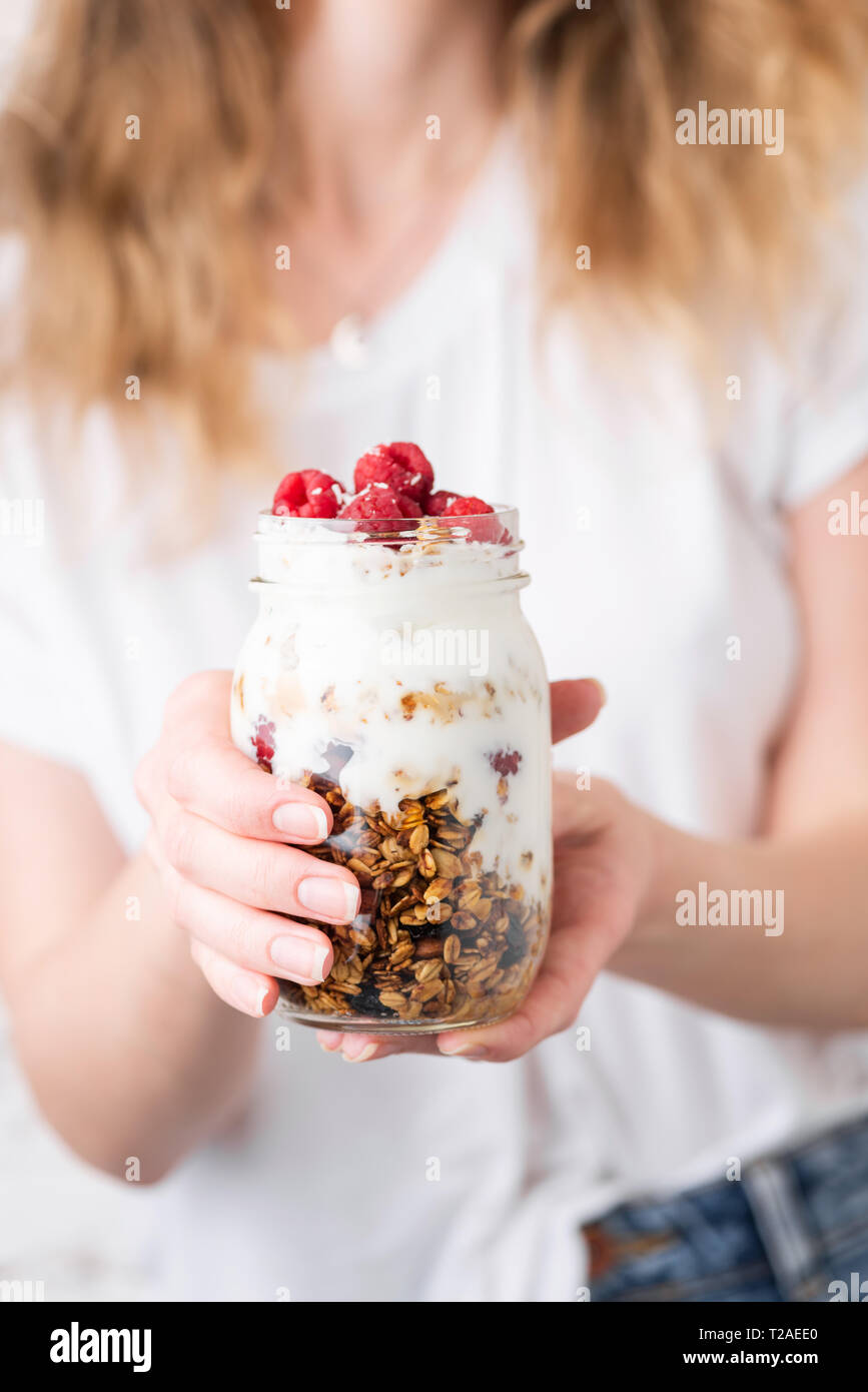 Le yogourt avec granola et les petits fruits dans le bocal de femmes. Femme blonde en chemise blanche holding pot de granola avec parfait au yogourt et petits fruits dans les mains. H Banque D'Images