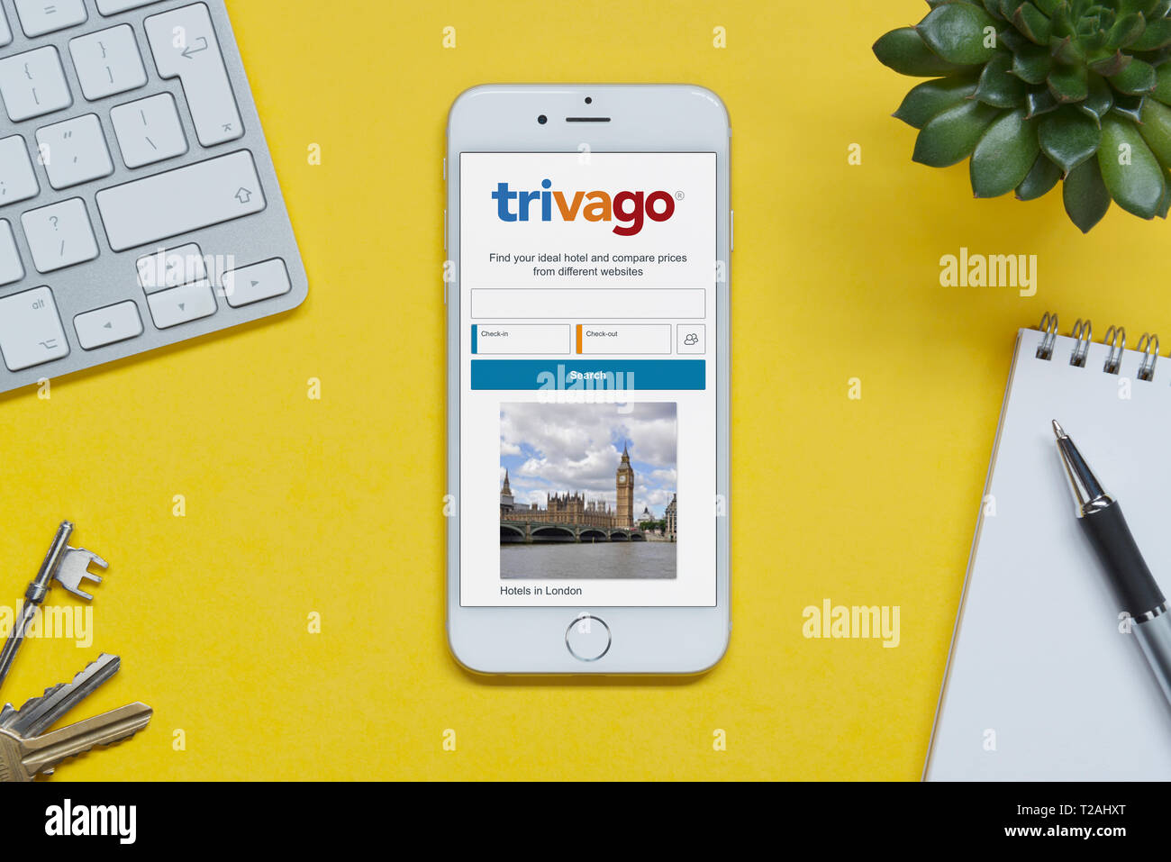 Un iPhone montrant le site Trivago repose sur un fond jaune table avec un clavier, les touches, le bloc-notes et des plantes (usage éditorial uniquement). Banque D'Images