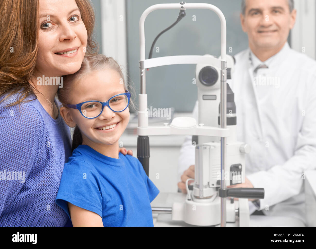 Jolie fille en bleu verres posant dans la clinique d'ophtalmologie médicale avec la mère. Médecin spécialiste de l'œil à maturité, smiling at camera, pa consulting Banque D'Images