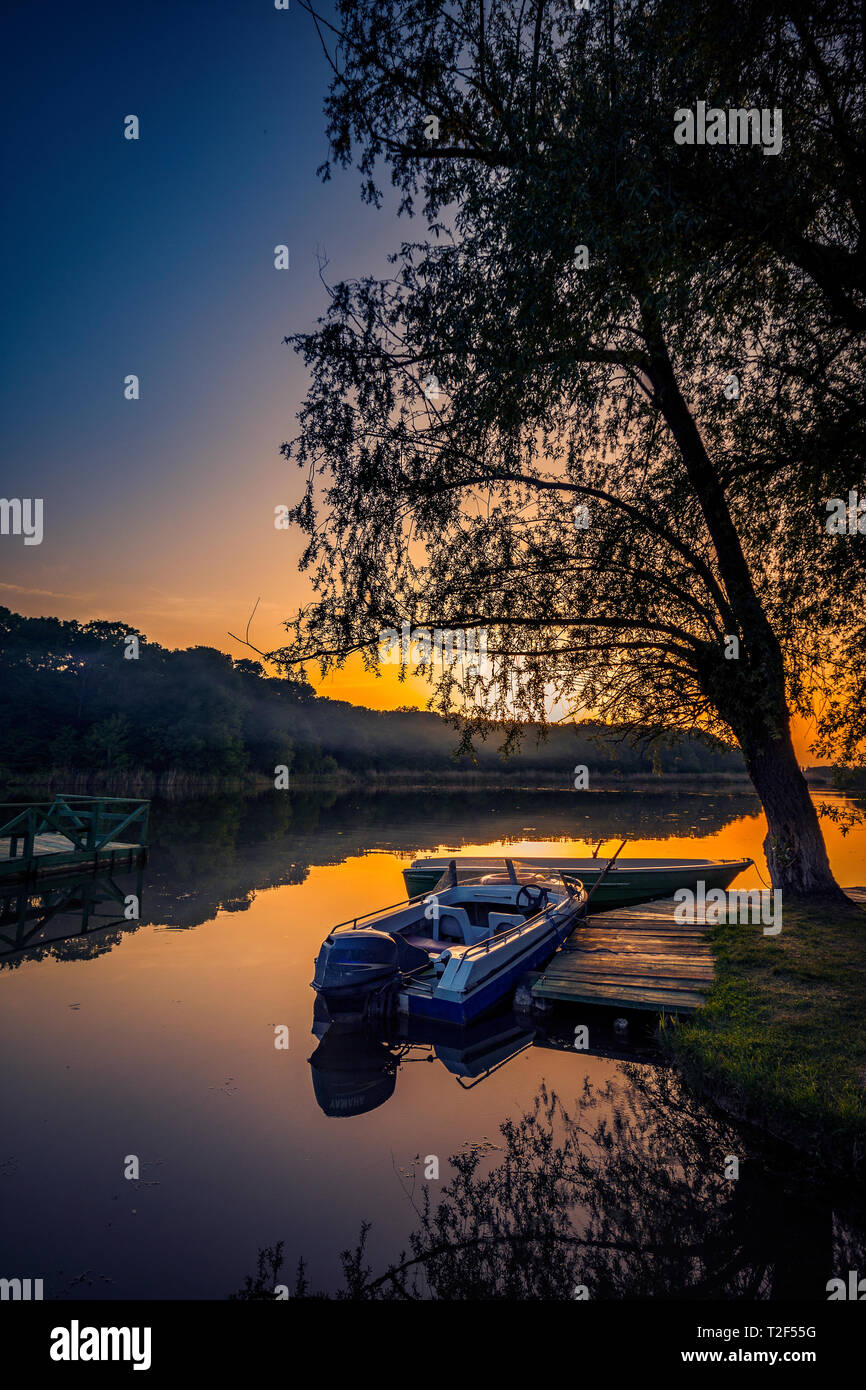 Bateaux de pêche sur un lac près d'un saule et d'un ponton au coucher du soleil contre un ciel clair Banque D'Images