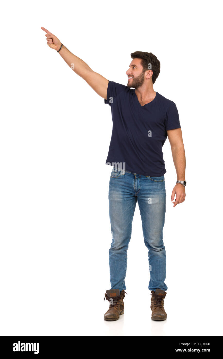 Bel homme en jeans, bottes et t-shirt bleu est debout, pointant vers le haut et en montrant quelque chose. Studio pleine longueur shot isolé sur blanc. Banque D'Images