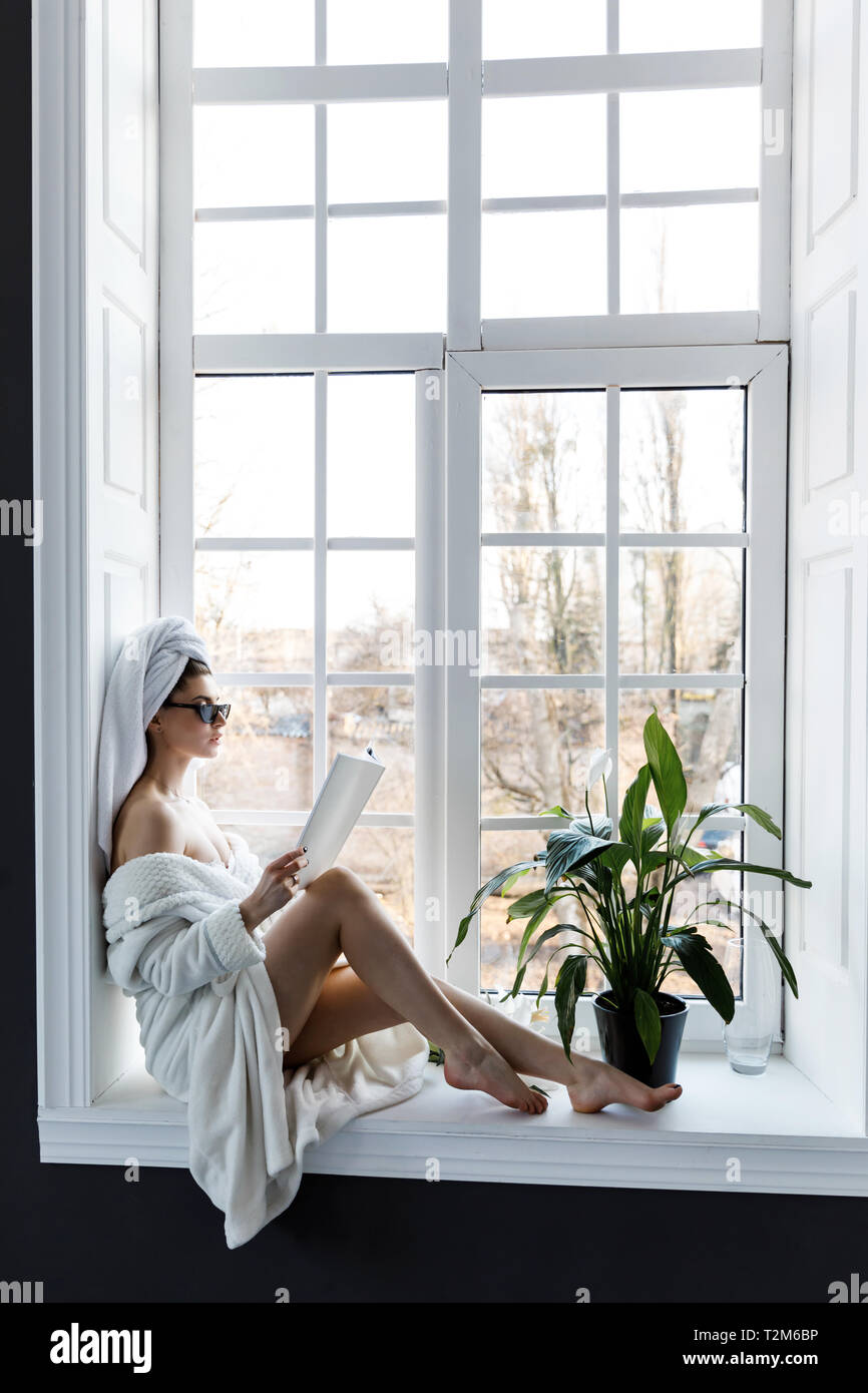 Une jeune femme vêtue d'une robe blanche et avec des serviettes sur la tête assis sur le rebord de la fenêtre et la lecture d'un magazine. Lieu de loisirs Banque D'Images