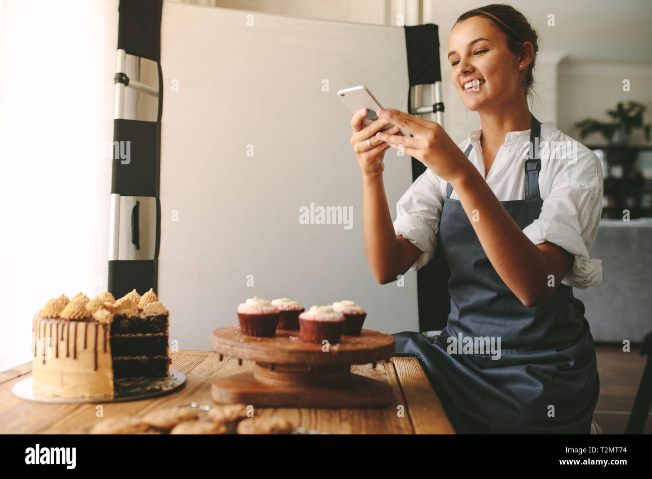 Happy woman chef de prendre une photo de pâtisseries sur une planche en bois avec smart phone alors que debout à la cuisine. Baker femmes prendre des photos de la de Banque D'Images