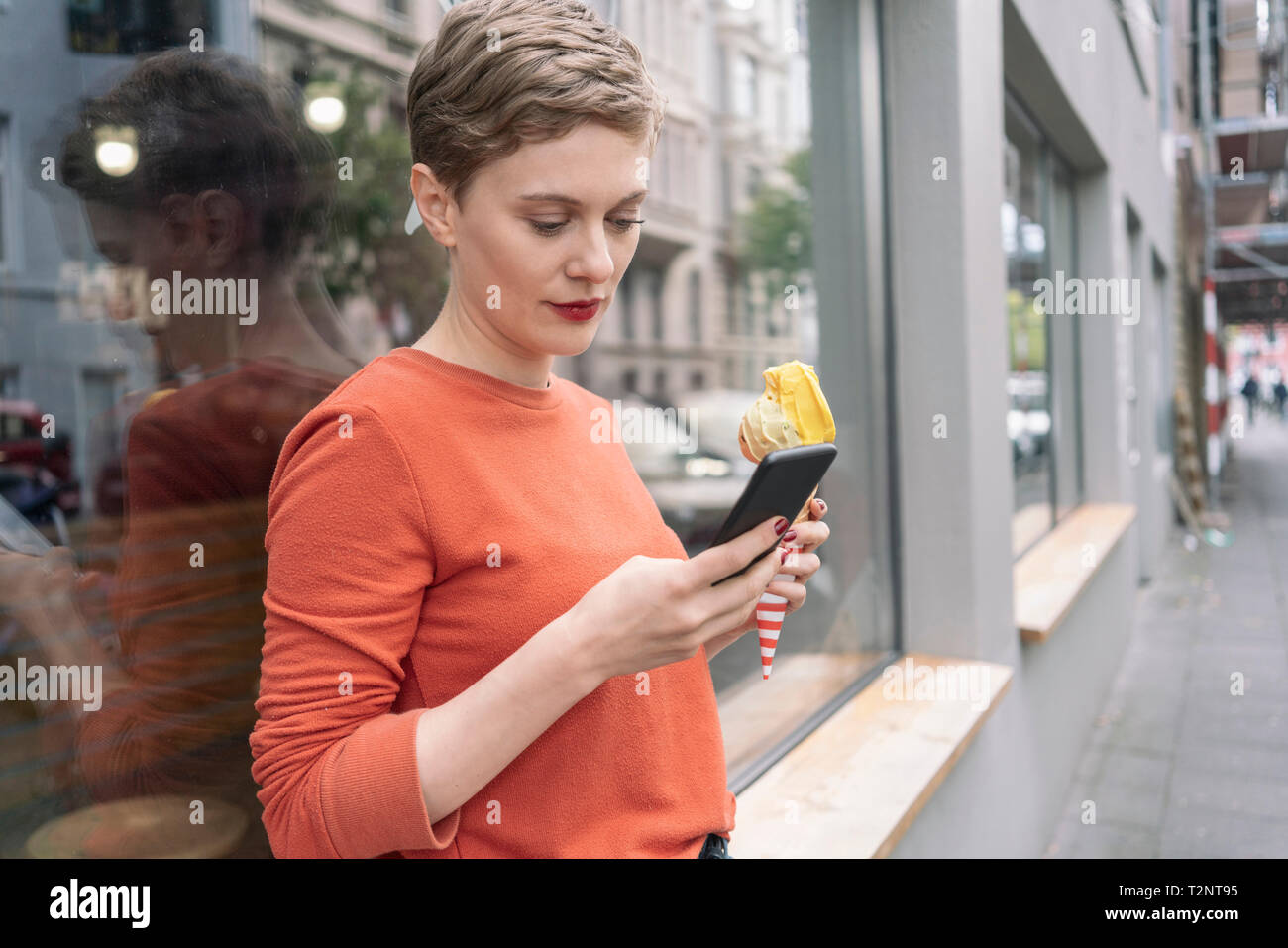 Woman holding ice cream et l'utilisation de smartphone à l'avant du magasin, Cologne, Nordrhein-Westfalen, Allemagne Banque D'Images