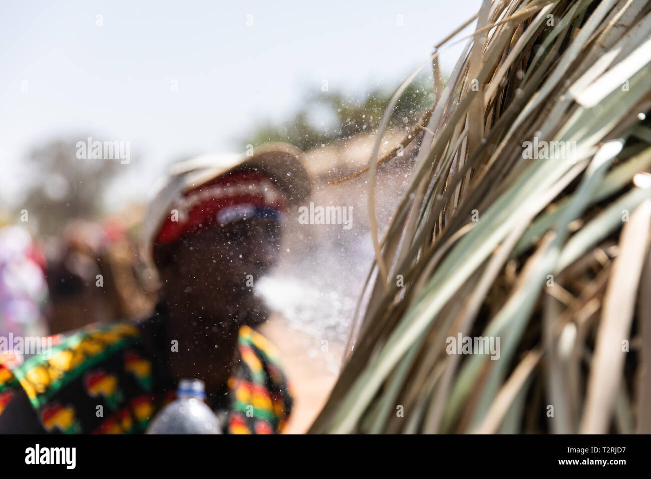 JANJANBUREH, la Gambie (19/01/2019) - Un maître de la Kompo Kankurang pulvérise de l'eau pour refroidir l'individu derrière la mascarade au cours de chaleur atteignant plus de 35 degrés celsius. Le Festival Janjanbureh Kankurang fait partie d'efforts conjoints de l'UE au Fonds d'affectation spéciale d'urgence pour l'Afrique et le projet d'autonomisation des jeunes Gambiens pour stimuler le tourisme à Janjanbureh. En préservant et en célébrant le patrimoine culturel de l'Kankurang, c'est la création de nouvelles possibilités économiques pour les jeunes Gambiens sous la forme d'une augmentation du tourisme. Banque D'Images