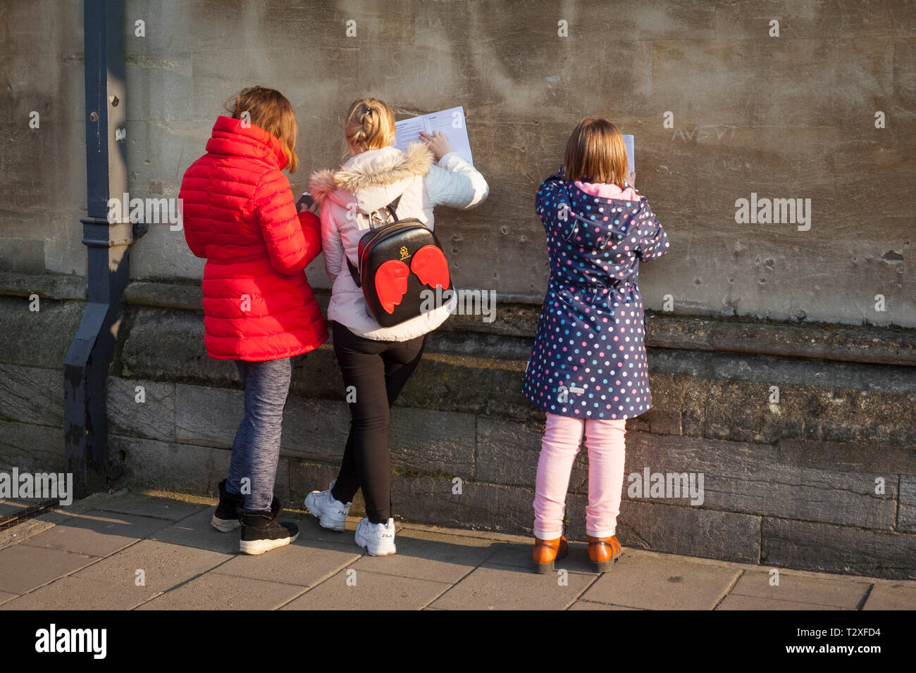 Les enfants de l'école sur une journée à Oxford en vêtements aux couleurs vives de remplir leur tournée quiz réponses appuyé contre un mur en pierre ancienne Banque D'Images
