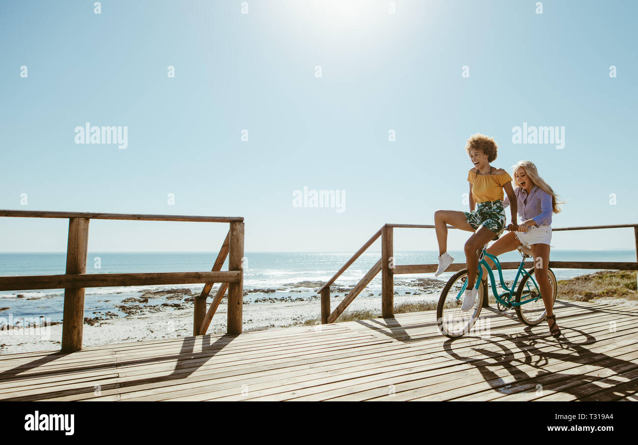 Les jeunes femmes joyeuse de la bicyclette ensemble. Les meilleurs amis s'amusant sur un vélo à la station de Boardwalk. Banque D'Images