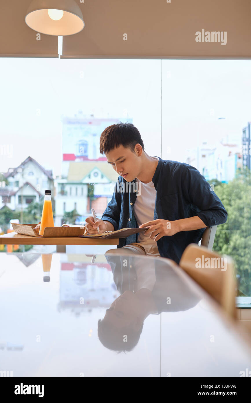 Beau jeune designer graphique créatif écrit des notes et croquis dans l'ordinateur portable tout en étant assis à table en bois à l'intérieur du café. L'homme réfléchi w Banque D'Images