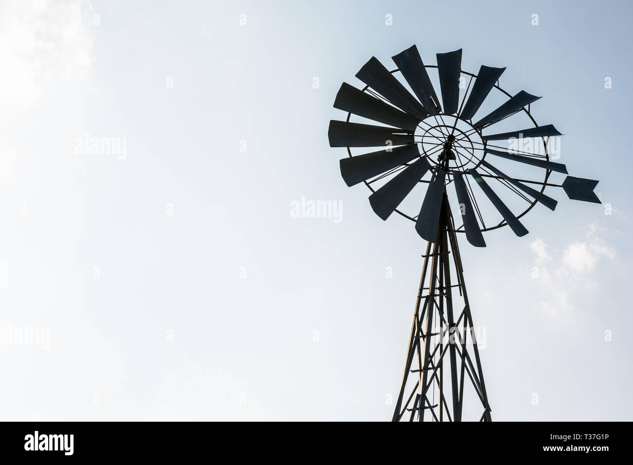 Low angle view of an old-fashioned, multi-lames, metal pompe éolienne au sommet d'un pylône en treillis en rétro-éclairage contre un ciel bleu pâle. Banque D'Images