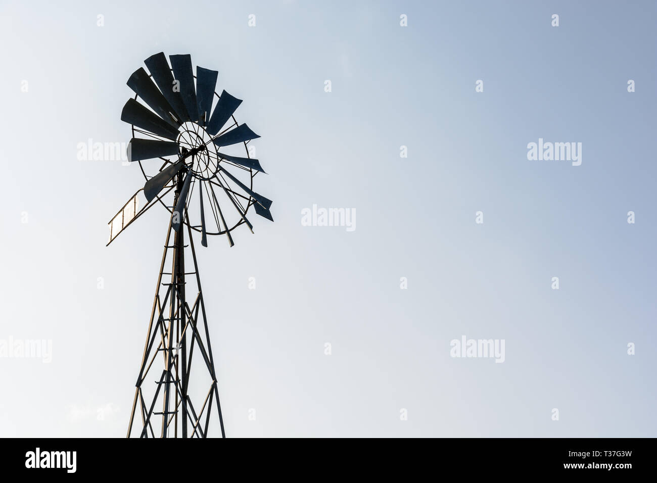 Low angle view of an old-fashioned, multi-lames, metal pompe éolienne au sommet d'un pylône en treillis en rétro-éclairage contre un ciel bleu pâle. Banque D'Images