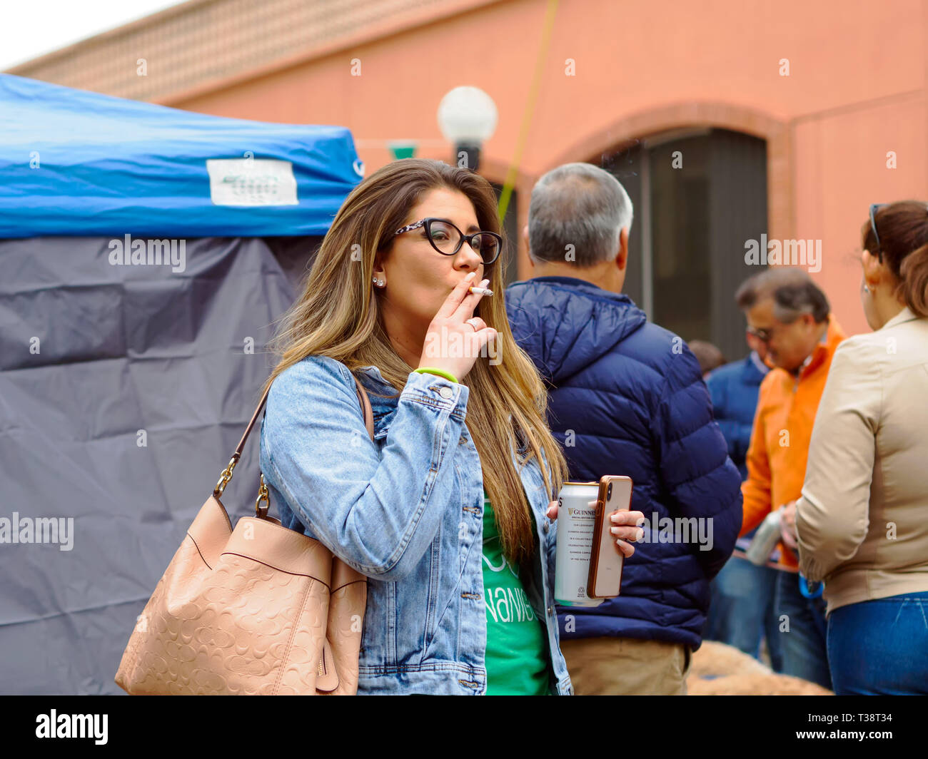 Une femme brune à lunettes et veste en jean bouffées d'une cigarette tout en tenant un téléphone cellulaire et peut de Guinness dans l'autre main. Banque D'Images