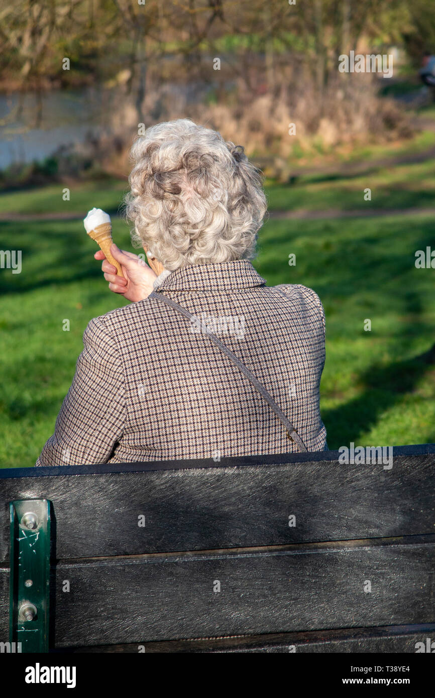 Une dame aux cheveux gris glamour jouit d'une glace sur un banc de parc Banque D'Images