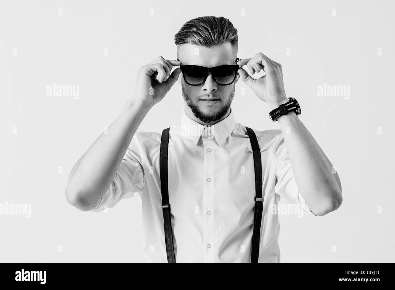 Tourné en noir et blanc de l'homme élégant et élégant avec bretelles chemise en portant des lunettes de soleil en position debout contre l'arrière-plan blanc. Je l'homme à la mode Banque D'Images