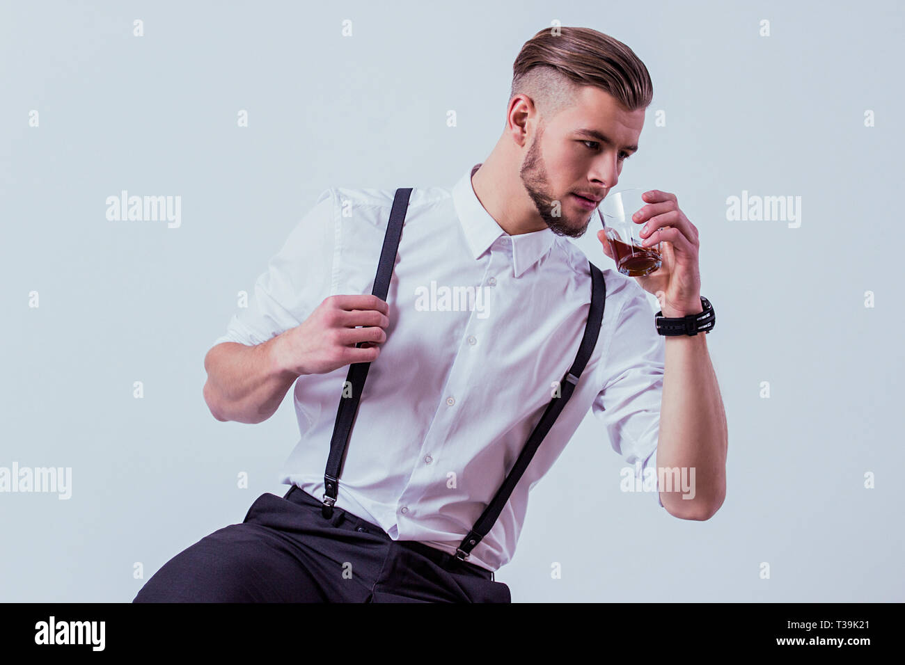 Studio shot of beau barbu en chemise blanche et des bretelles de boire de  l'alcool alors qu'il était assis à l'arrière-plan gris. Homme élégant à la  WEA Photo Stock - Alamy