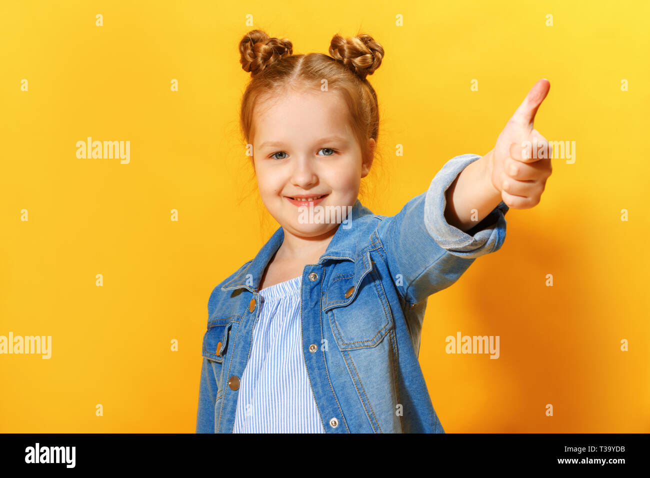 Closeup portrait of a cute joli petit enfant girl showing thumb up sur fond jaune. Banque D'Images