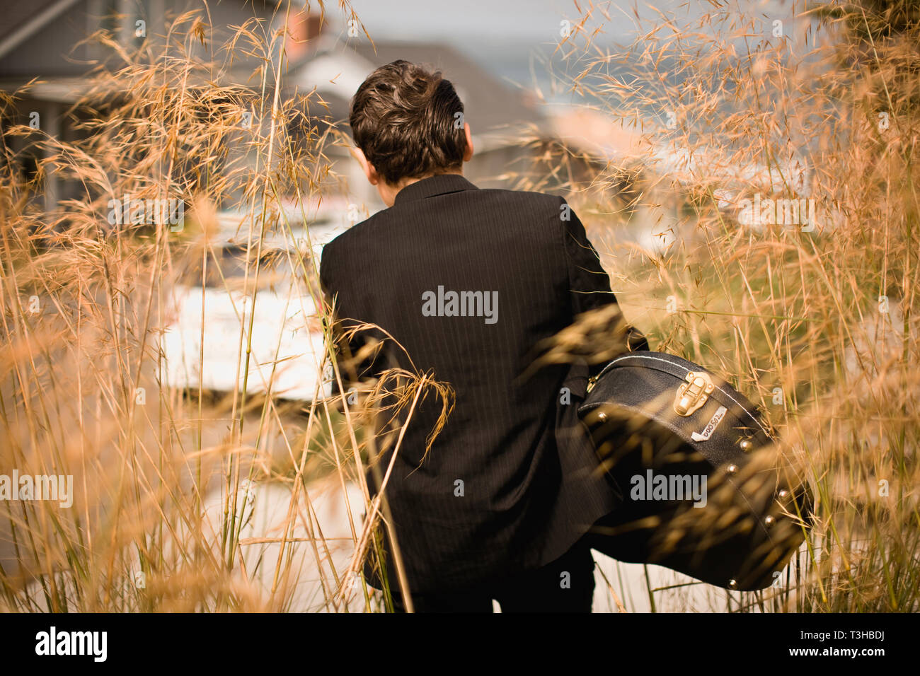 Garçon dans un costume et portant une guitare acoustique cas marche dans une colline de banlieue, dans les longues herbes miteux. Banque D'Images