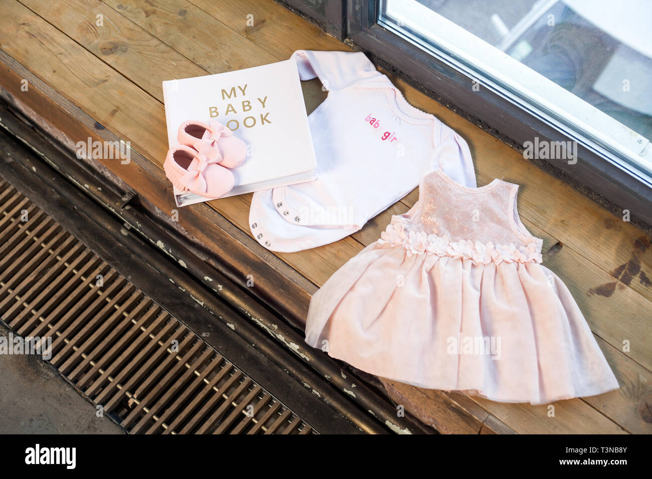 Rose robe vêtements bébé fille nouveau-né avec l'album livre, du corps et de petites chaussures sur fond de caisse en bois de la fenêtre. Banque D'Images