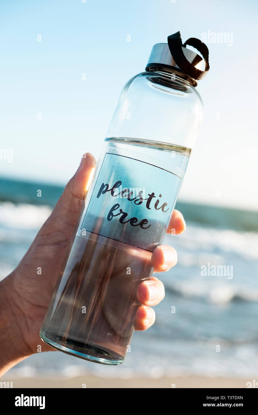 Libre d'un homme de race blanche tenant une bouteille d'eau réutilisable en verre avec le texte libre en plastique écrit en C, sur la plage, avec l'océan dans le background Banque D'Images