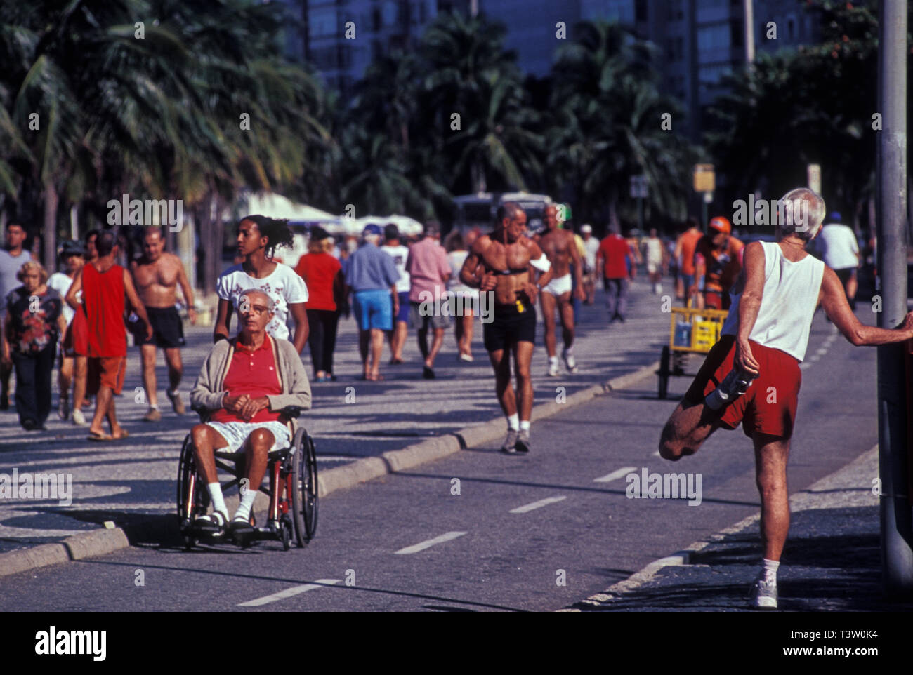 Contraste - plusieurs personnes en bonne santé les gens exercent près d'un utilisateur de fauteuil roulant handicapé homme âgé soigné par une jeune femme noire au trottoir de la plage de Copacabana, Rio de Janeiro, Brésil. Banque D'Images