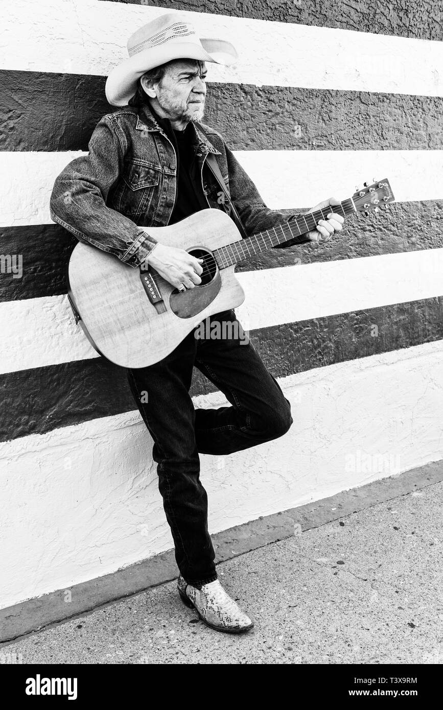 Old cowboy avec chapeau de cowboy et barbe grise joue une guitare tout en se tenant à côté d'un mur à rayures blanches et noires. Banque D'Images