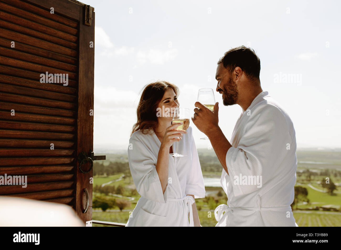 Sourit l'homme et de la femme tenant un verre de vin et de parler les uns aux autres. Couple en peignoir debout dans balcon de chambre bénéficiant d''un verre de vin. Banque D'Images