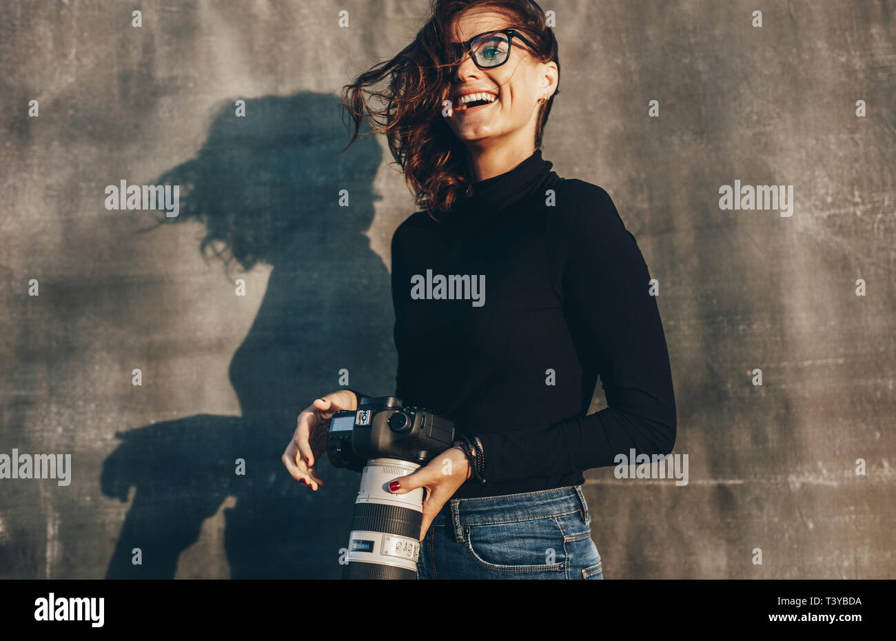 Laughing young woman holding un appareil photo DSLR contre fond brun. Femme photographe réussie sur un shoot photo. Banque D'Images