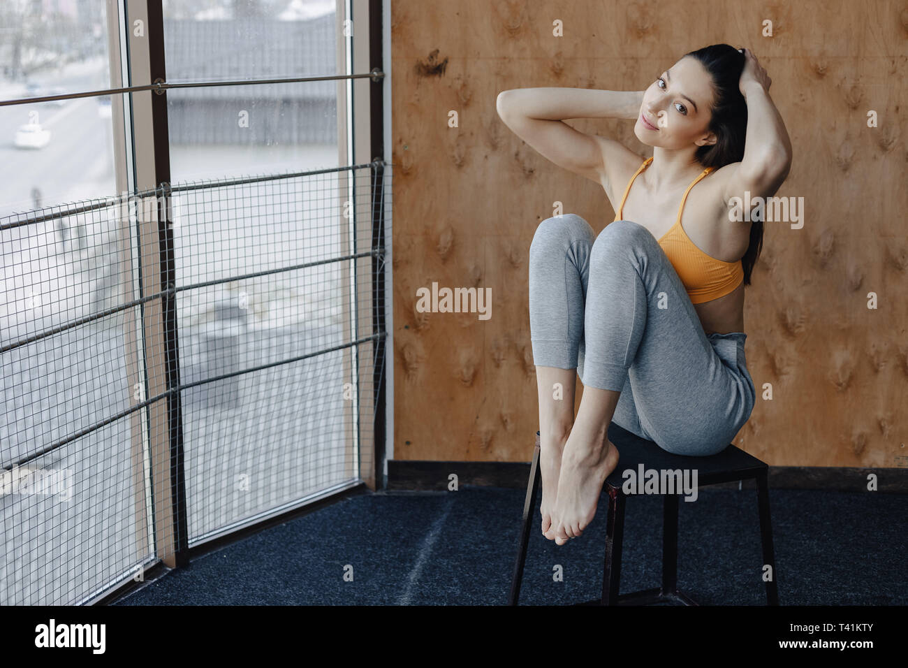 Young attractive fitness petite fille assis sur une chaise près de la fenêtre sur l'arrière-plan d'un mur en bois, reposant sur des cours de yoga à gym Banque D'Images