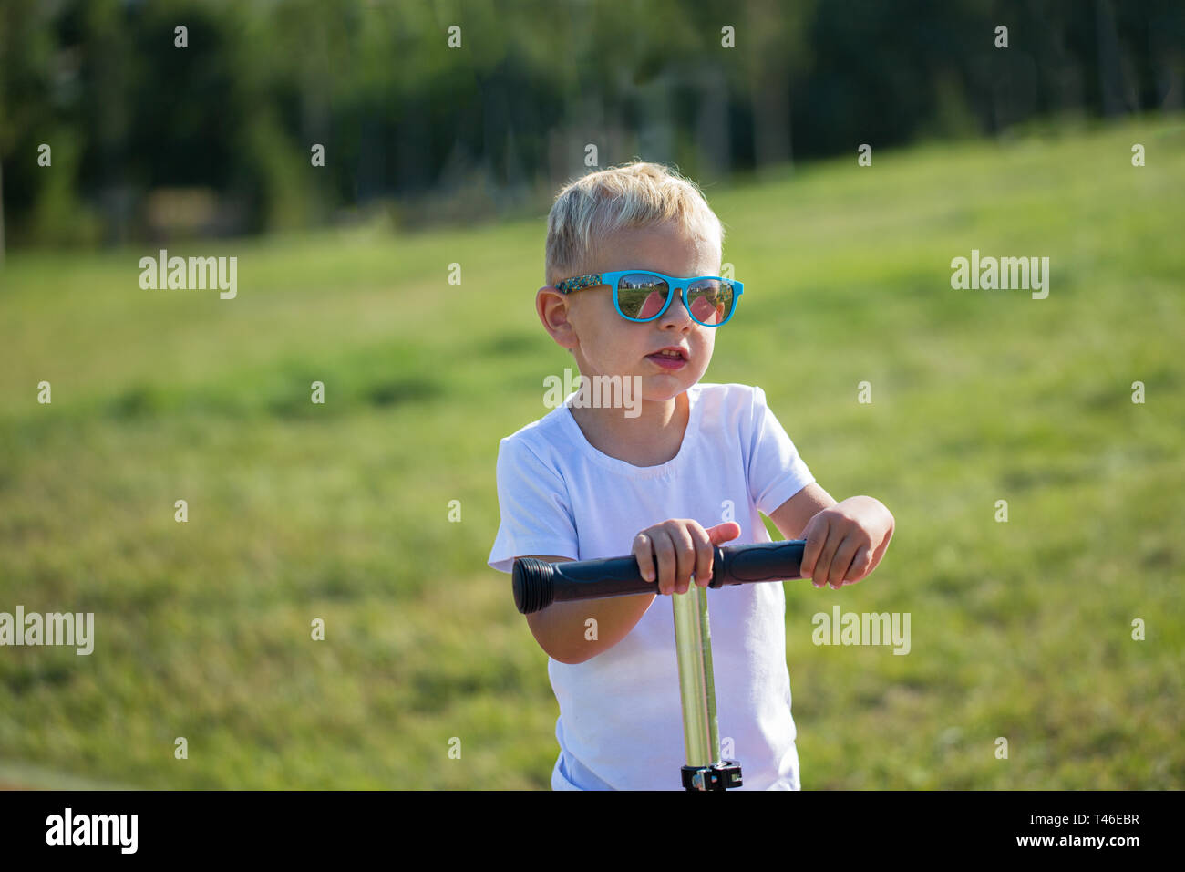 Beau petit Garçon à lunettes élégant monte un scooter sur une piste dans le parc sur une journée ensoleillée Banque D'Images